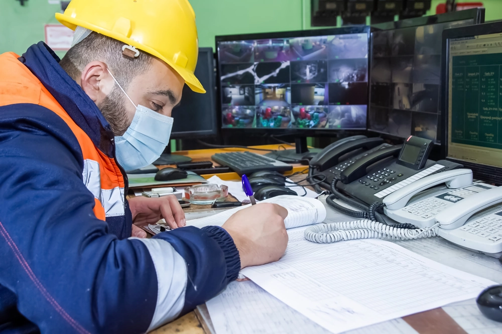 BHP-owiec w kasku i kamizelce zapisuje dokumentację nadzoru bhp przed monitorami CCTV
